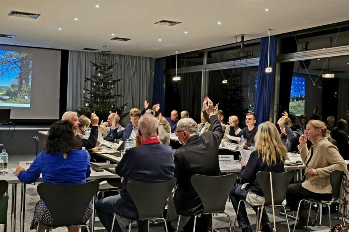 Personengruppe sitzt an Tischen in einem Raum mit Weihnachtsbaum und hebt zum Abstimmen die Hände