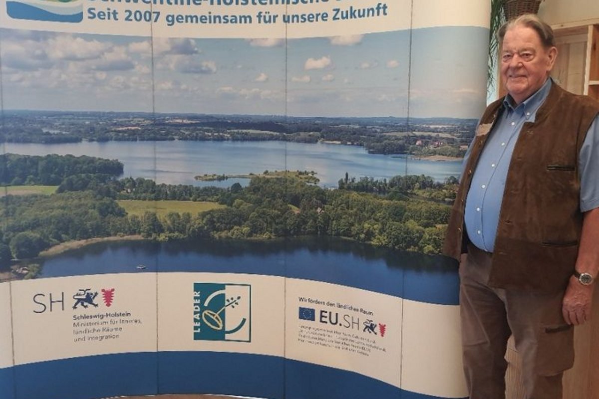 Joachim Schmidt steht neben einer gebogenen Plakatwand mit Landschaftsfoto und Logos der LAG AktivRegion Schwentine-Holsteinische Schweiz