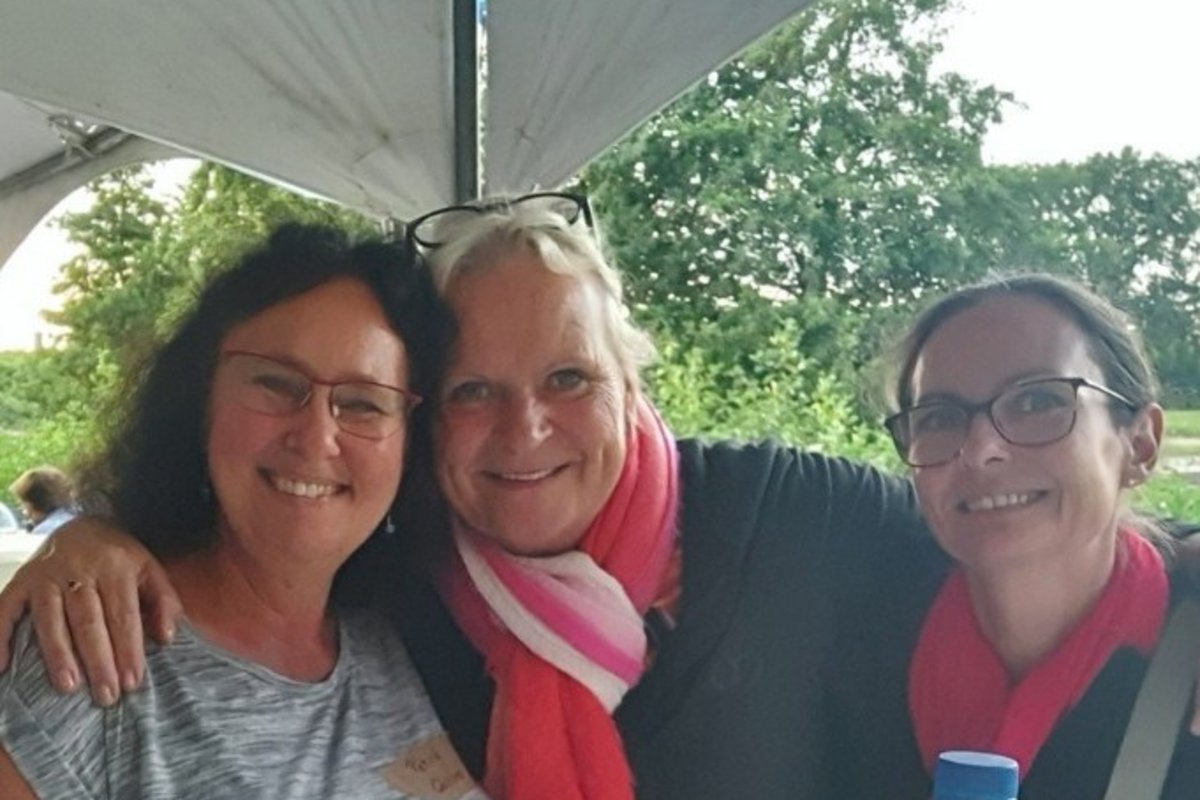 Drei lächelnde Frauen sitzen eng beieinander unter einem Zelt mit Bäumen im Hintergrund