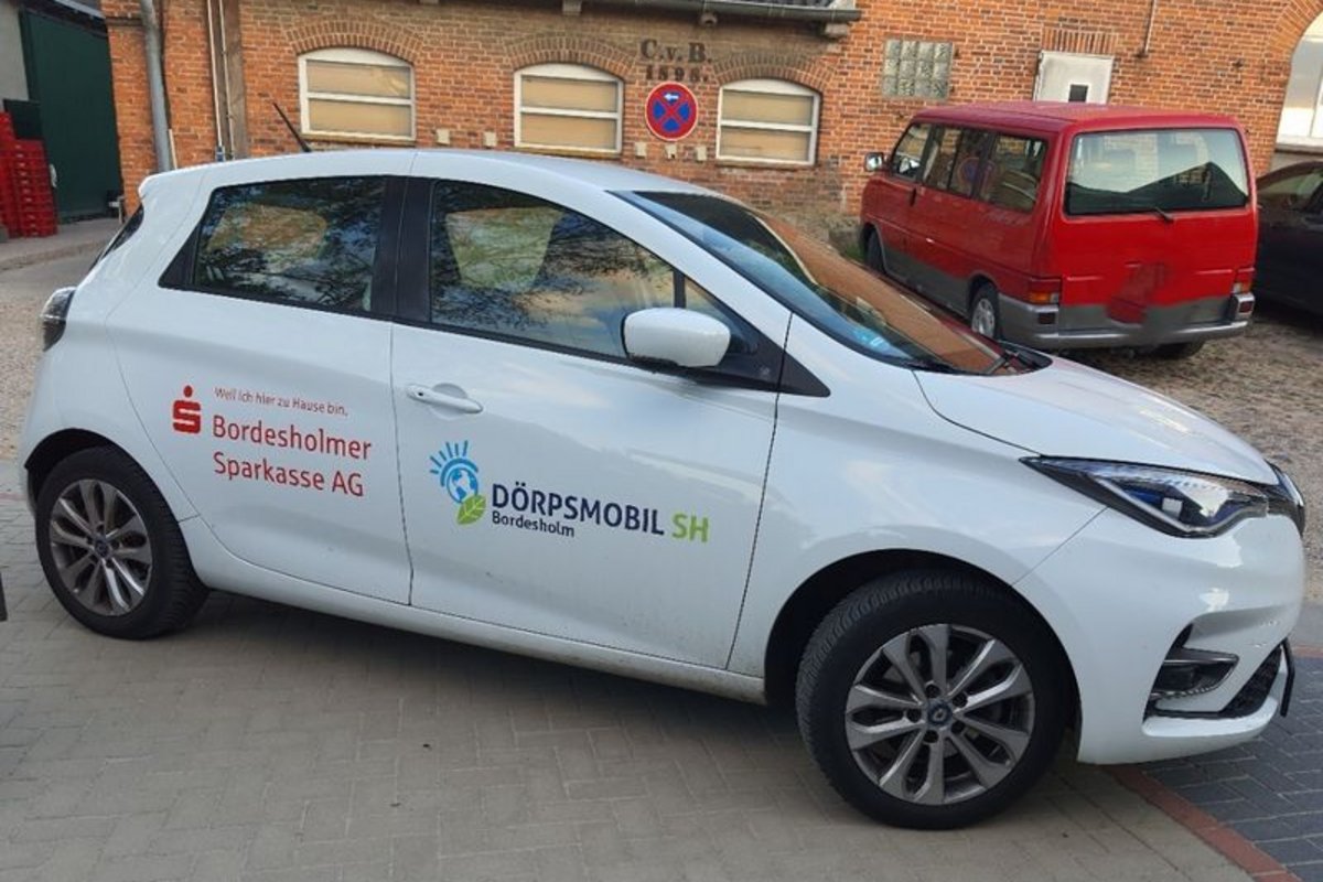 Weißes Auto mit Aufschrift "Bordesholmer Sparkasse AG" und "Dörpsmobil SH Bordesholm" steht vor einem Backsteingebäude