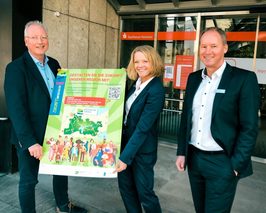 ein Mann und eine Frau halten gemeinsam ein Plakat, während ein lächelnder Mann rechts daneben steht